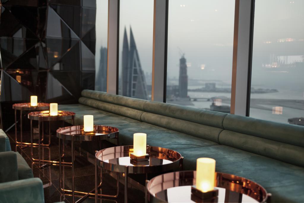 The Domain Bahrain Hotel And Spa - Adults Friendly 16 Years Plus Manama Luaran gambar The photo depicts a stylish lounge area with a modern aesthetic. There are several circular tables made of metallic materials, each topped with a small glowing candle. A long, plush seating area in a teal color runs along the window, offering a view 